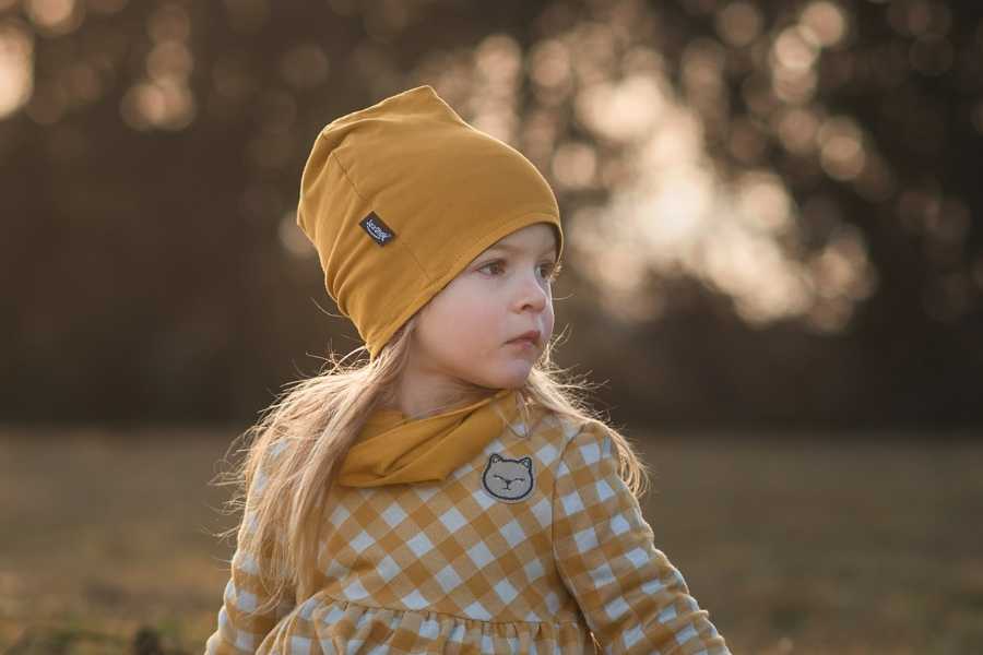 Dievčatko s károvanými šatami a horčicovo žltou čiapkou a nákrčníkom sedí pri západe slnka na lúke a vážne hľadí do diaľky